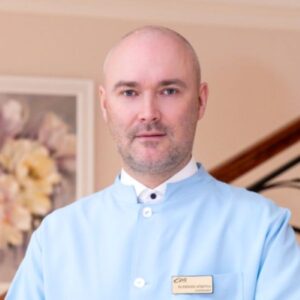 Elite Clinic 2 Dr. Deniss Sõritsa in a light blue medical coat with a name badge, standing indoors with a floral painting in the background.