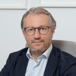 Dr. Gkaravelas from Ovagenesis is sitting in a black blazer and a blue shirt, wearing glasses, on a gray armchair in his office.