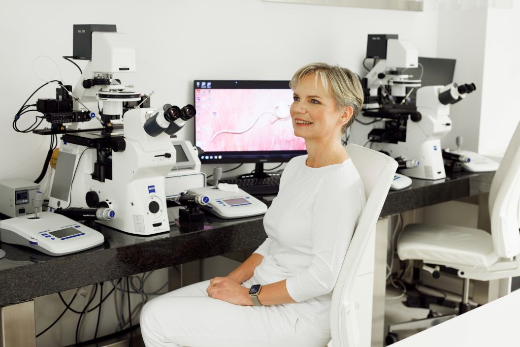 MSc Renata Hüttelova in an IVF laboratory, seated next to microscopes and fertility testing equipment, Zeiss-branded devices visible on the desk.