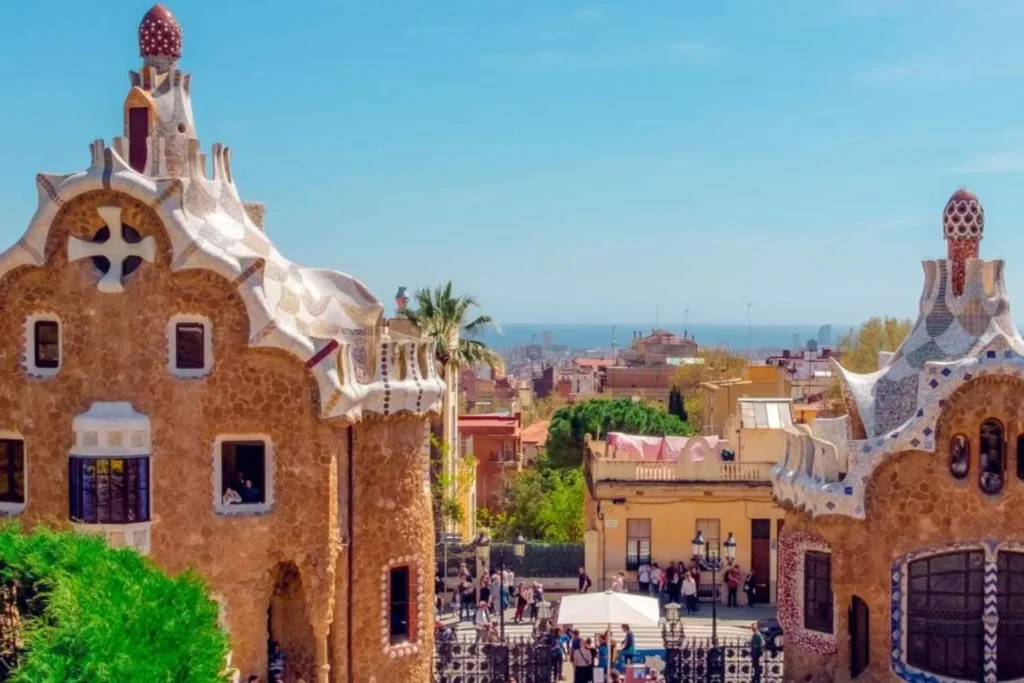IVF Spain Reviews: What Patients Say About Fertility Treatments in Spain 7 Two stone buildings with curved, white mosaic roofs stand on either side of a walkway, with people gathered below and a city and blue sea visible in the background under a clear sky.